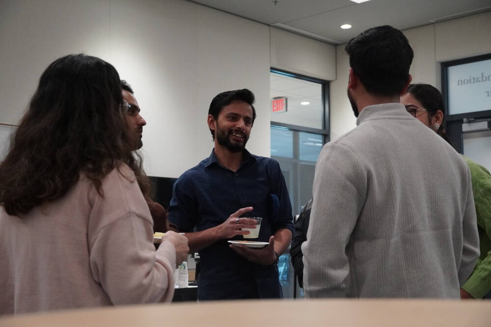 A group of people engage in a conversation at an indoor space. The person in the center, holding a drink and a plate, smiles while speaking with others gathered around.